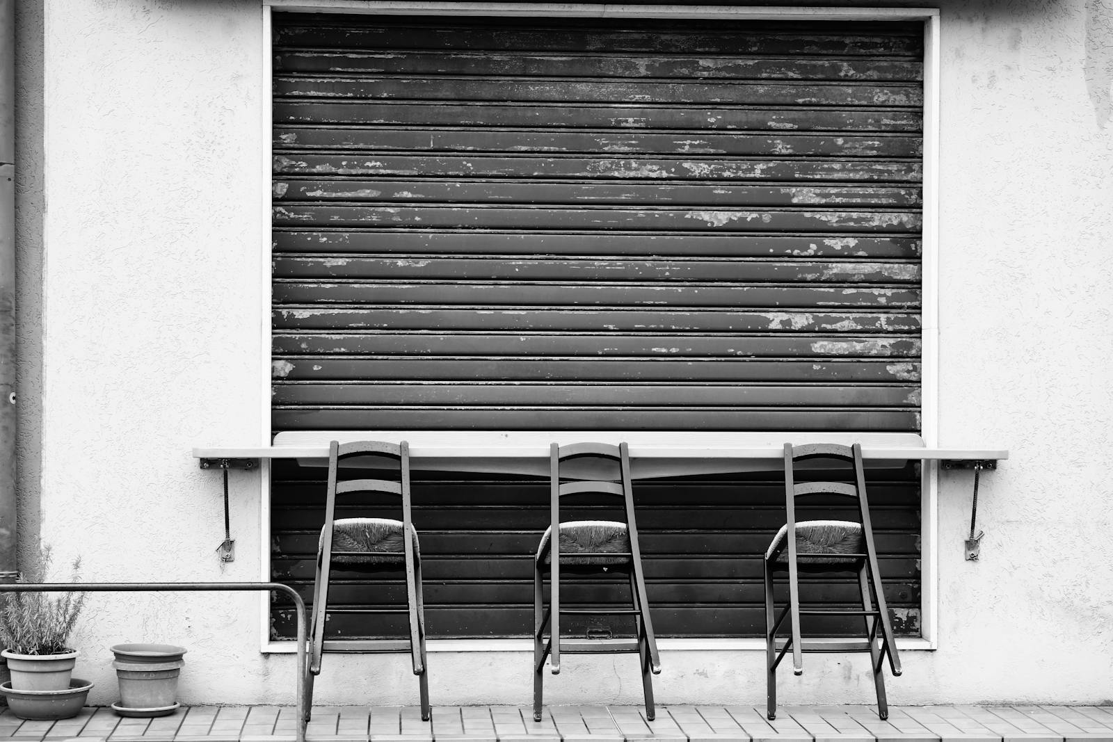 A black and white image of an outdoor cafe with three chairs in front of a closed shutter.